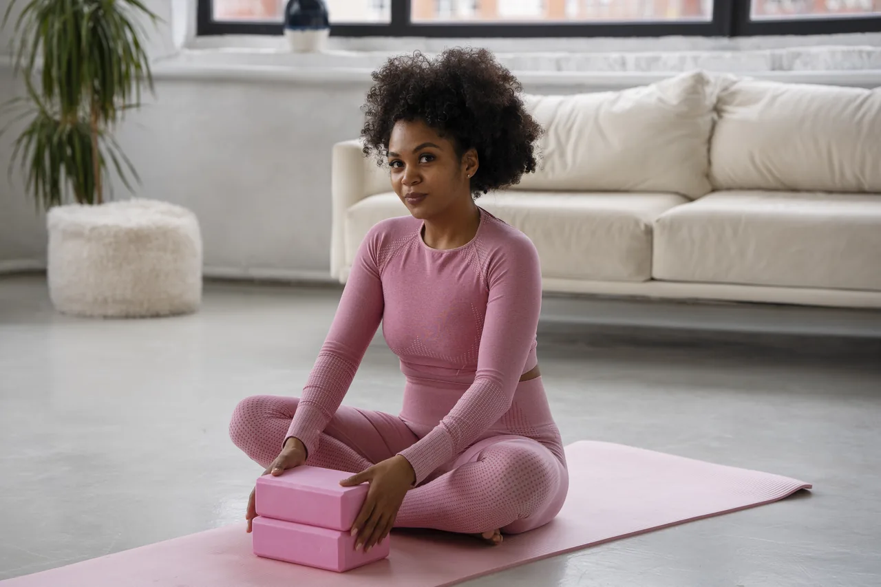 a black pregnant woman sitting on a pink mat with a yoga block
