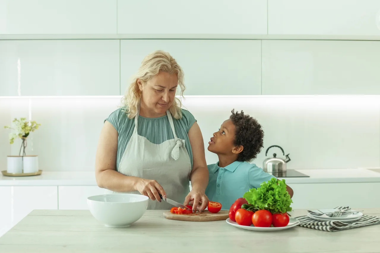 a woman in her fifties chopping vegetables while a small boy leans in talking to her