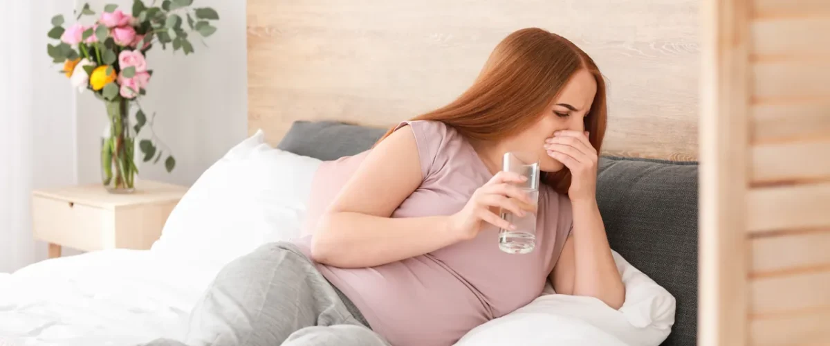 a pregnant woman half-lying in bed with one hand over her mouth and a glass of water in the other