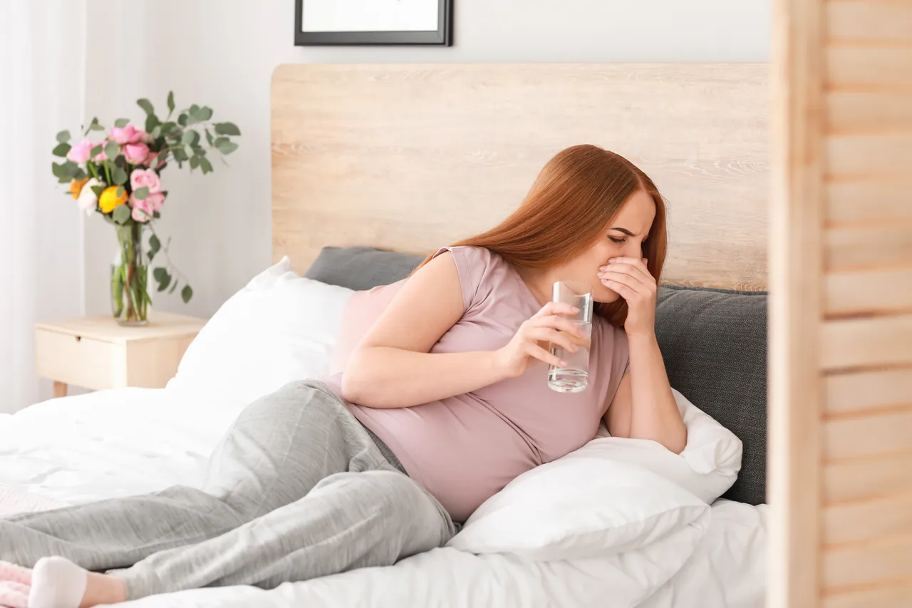 eine Schwangere liegt halb im Bett, eine Hand vor dem Mund, in der anderen ein Glas Wasser