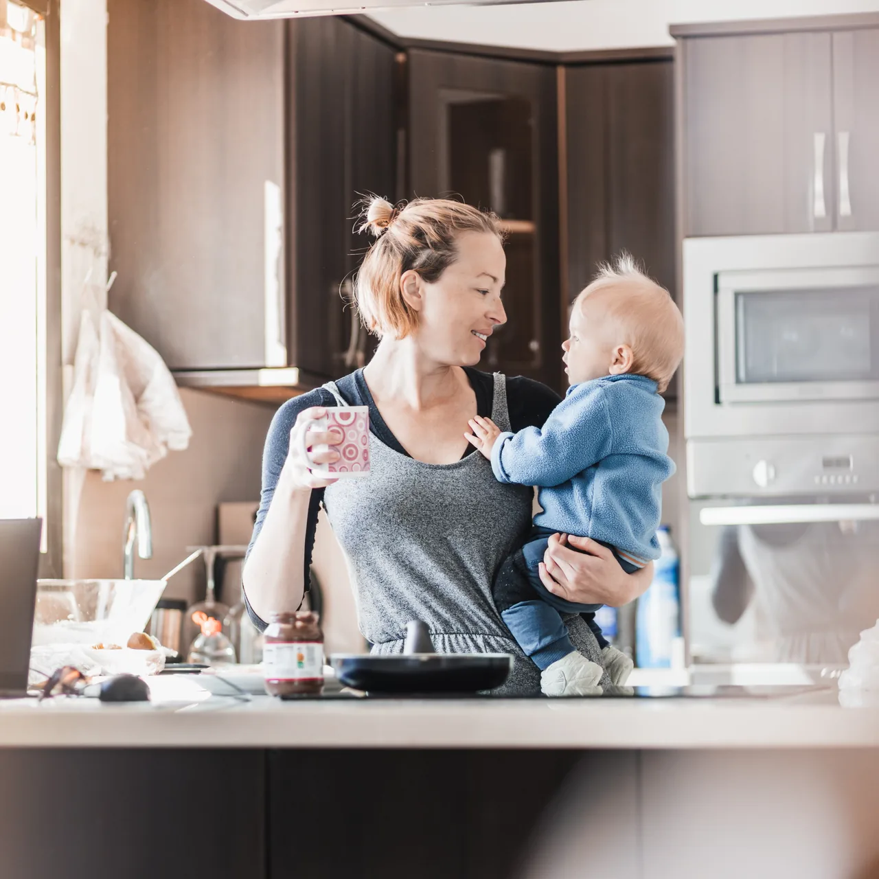 a woman holding a mug and carrying her baby on one hip in a cluttered everyday kitchen