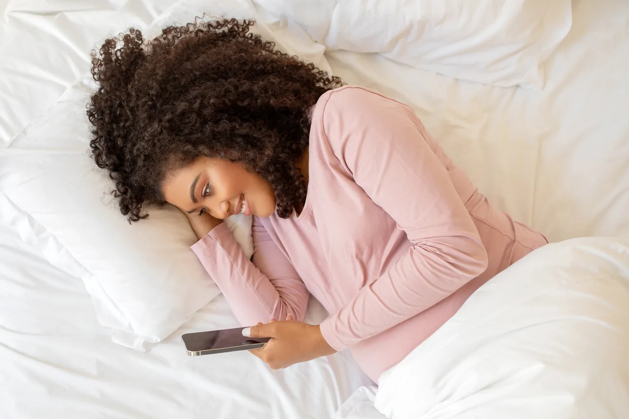eine Schwangere liegt im rosa Oberteil seitlich im Bett und liest auf ihrem Telefon
