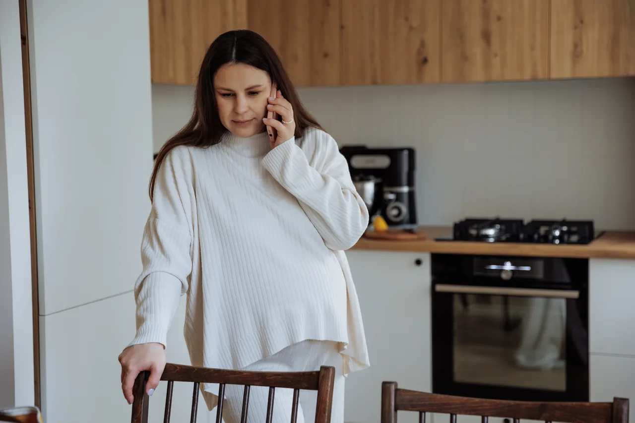 eine Schwangere im weiten Strickpullover telefoniert in ihrer Küche