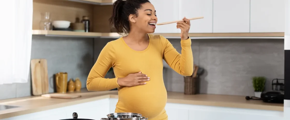 a pregnant woman tasting from a wooden spoon at the hob with one hand on her belly