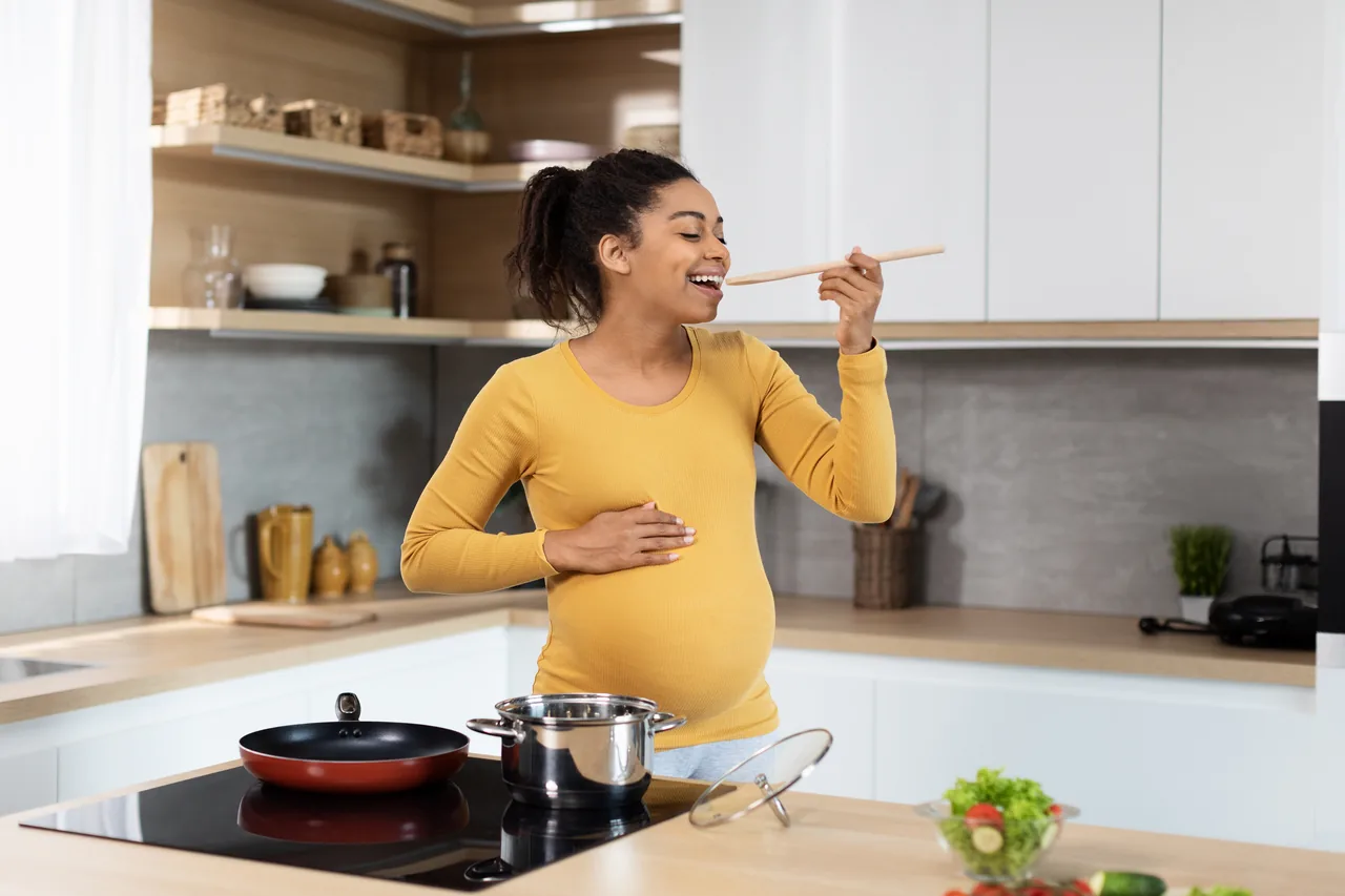 uma grávida prova de uma colher de pau ao fogão com uma mão na barriga