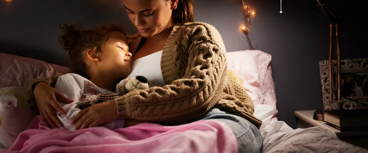 a woman and her small daughter asleep together in bed under a string of fairy lights