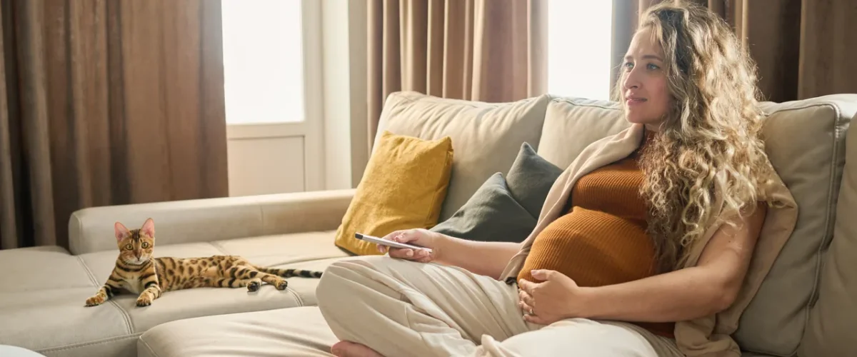 a pregnant woman sitting cross-legged on a sofa with her phone while the cat lies beside her