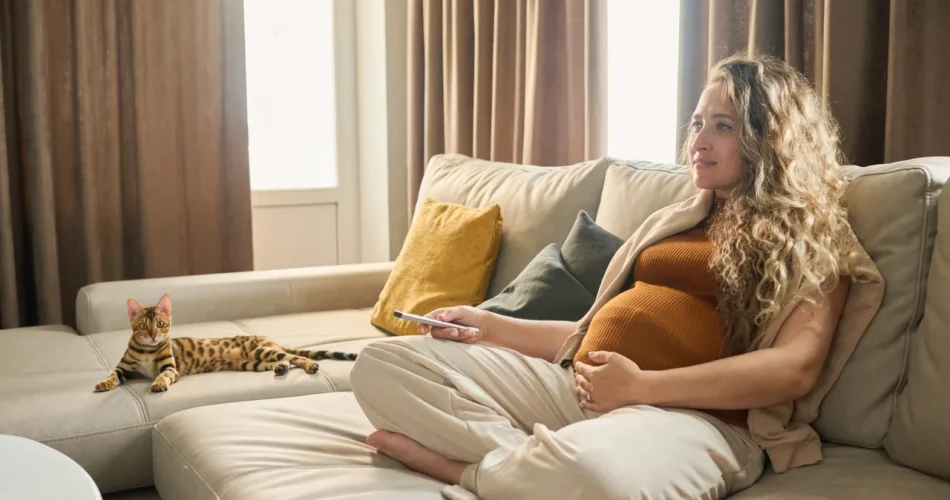 a pregnant woman sitting cross-legged on a sofa with her phone while the cat lies beside her