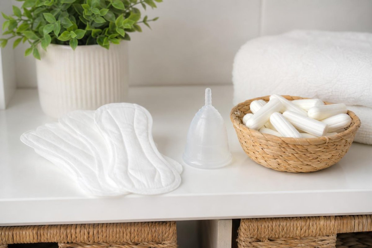 Weiße Binden, eine durchsichtige Menstruationstasse und ein Korb mit Tampons auf einem Regal neben Handtuch und Pflanze